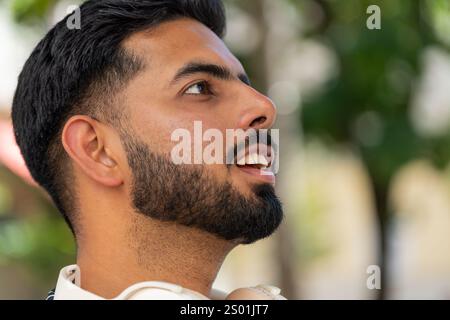 Giovane e premuroso turista maschile con la barba e cuffie wireless intorno al collo che guardano in alto e sorridono. Vista laterale del viaggiatore indiano felice e positivo che si trova sul parco cittadino. Stili di vita della città. Foto Stock