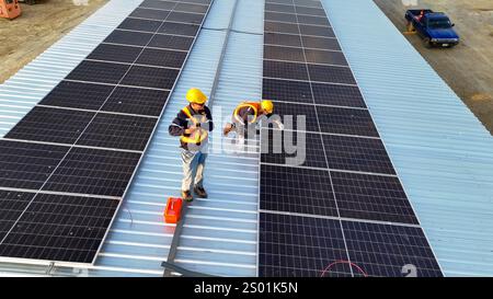 Due operai edili stanno installando pannelli solari su un tetto in metallo indossando caschi e giubbotti di sicurezza. Il sole luminoso illumina la scena A. Foto Stock