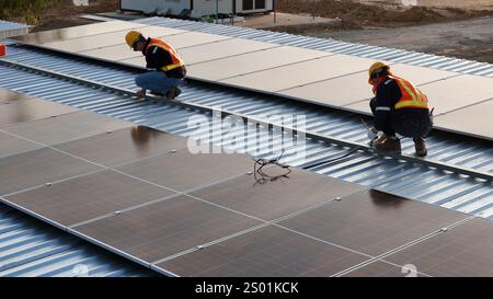 Due lavoratori che indossano caschi e giubbotti di sicurezza proteggono i pannelli solari su un tetto metallico. Il cantiere è esposto alla luce solare e mostra energia Foto Stock
