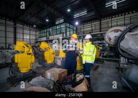 Due ingegneri discutono i dettagli operativi mentre esaminano i bracci robotici in un magazzino ben illuminato. L'attenzione è rivolta al lavoro di squadra e all'integrazione tecnologica in Foto Stock