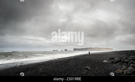 Paesaggio spettacolare che mostra un turista che cammina sulla spiaggia di sabbia nera di reynisfjara, islanda, con colonne di basalto, scogliere e tempo burrascoso Foto Stock