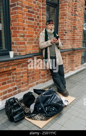 Ritratto verticale a tutta lunghezza di un anziano senzatetto con barba bianca che usa il telefono cellulare che guarda sullo schermo mentre si appoggia su un muro di mattoni nella strada della città Foto Stock