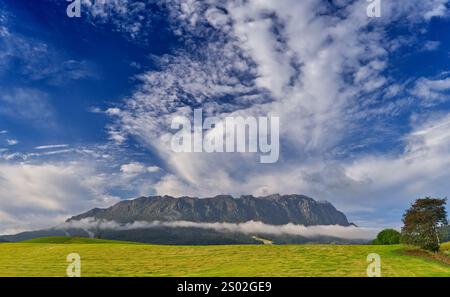 Vista della catena monolitica del Monte Roland con nebbia e nebbia al mattino e cielo azzurro sul verde pendio erboso, Sheffield, Tasmania, Australia Foto Stock