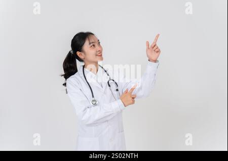 Una bella e positiva dottoressa asiatica in uniforme si erge su uno sfondo bianco di studio, sorridendo e puntando ad uno spazio vuoto, come se fosse re Foto Stock