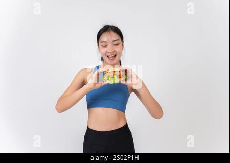 Una allegra donna asiatica in abbigliamento sportivo con un hamburger, in piedi su uno sfondo bianco dello studio. equilibrio tra cibo e allenamento, dieta, perdita di peso, l Foto Stock