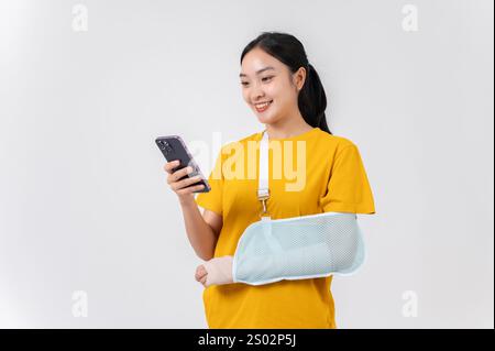Una donna asiatica sorridente e spensierata con il braccio rotto, avvolta in una bendatura in gesso e sostenuta da una fionda dopo una lesione accidentale, usando il suo smartph Foto Stock