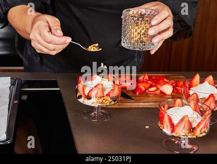 Un primo piano di granola cosparso in tazze di vetro con fragole appena affettate su un asse di legno in un ambiente di cucina. Foto Stock