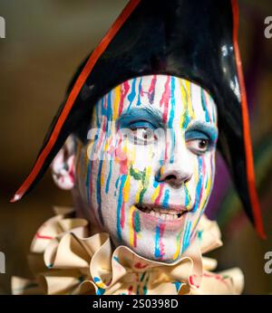 Una persona con un volto colorato e dipinto con strisce di blu, rosso e giallo, che indossa un grande cappello nero e un colletto con volant. Lo sfondo è B. Foto Stock