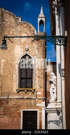 Un edificio storico con una facciata in mattoni e una finestra ornata, caratterizzato da una statua di una figura bianca sulla destra. La scena è ambientata contro un cielo blu W. Foto Stock