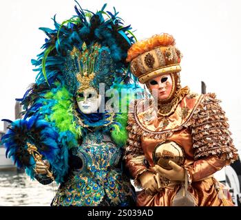 Due individui elegantemente vestiti in elaborati costumi e maschere, che mostrano colori vibranti e disegni intricati, in piedi accanto a un corpo d'acqua. Foto Stock
