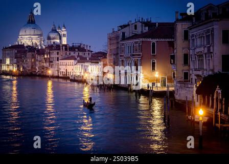 Una serena vista serale di un canale a Venezia, con gondole che scivolano attraverso l'acqua. La scena è illuminata da luci stradali calde che riflettono o Foto Stock