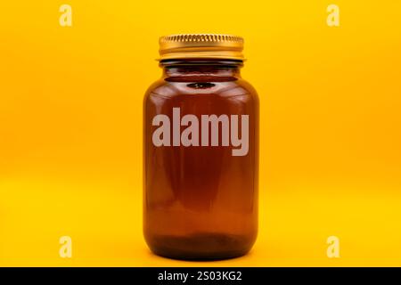 Primo piano di un vaso in vetro ambrato vuoto con coperchio dorato, isolato su uno sfondo giallo brillante, perfetto per il branding o il packaging. Foto Stock