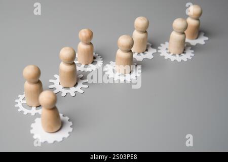 Un'immagine concettuale con figure di legno in piedi su ingranaggi interconnessi. Lavoro di squadra, collaborazione e la natura interconnessa del concetto di lavoro. Foto Stock