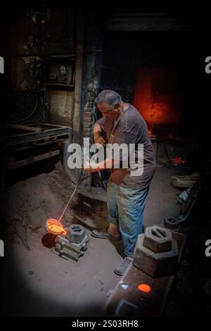 Un operaio di fonderia in un'officina ad Alessandria, Egitto, che fonde metallo fuso in un ambiente tradizionale. Un'immagine del fondatore dell'ottone riflette l'artigianato locale Foto Stock