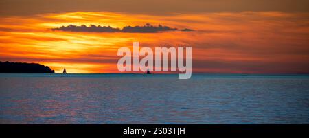 Un tramonto sereno su un mare calmo, con due barche a vela che si stagliano contro il vivace cielo arancione e blu. L'orizzonte è illuminato dolcemente, creando un peacefu Foto Stock