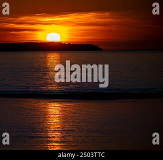 Uno splendido tramonto sulle acque calme, con il sole che si tuffa sotto l'orizzonte, che getta un riflesso dorato sulla superficie. Alberi sagomati allineano il dis Foto Stock