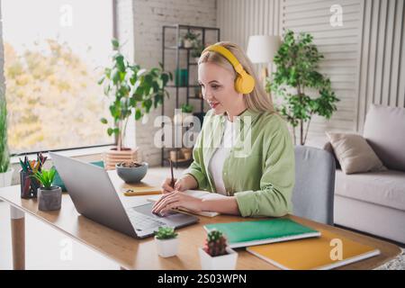 Giovane donna che indossa le cuffie gialle utilizzando un computer portatile e che scrive su un notebook a una scrivania in un ambiente accogliente e ben illuminato. Foto Stock