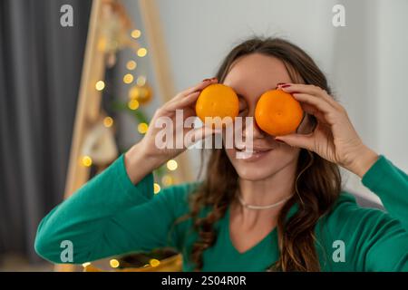 La donna copre scherzosamente gli occhi con due cachi maturi. Sorride, indossa un maglione verde. Un albero di Natale sfocato è sullo sfondo. Festività Foto Stock