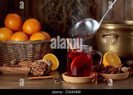 Il VIN brulé viene versato in una tazza di bicchiere. VIN brulé con ingredienti su un vecchio tavolo di legno. Foto Stock