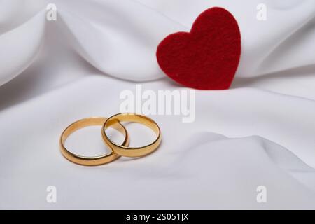 Fedi nuziali dorate e cuore rosso su sfondo bianco in tessuto Foto Stock