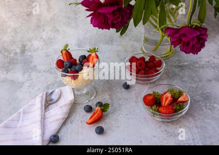 Dessert estivo fresco con gelato cremoso alla vaniglia guarnito con frutti di bosco maturi in una ciotola di vetro, accompagnato da peonie rosa su una superficie ruvida Foto Stock