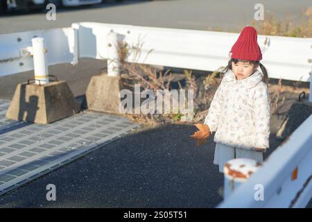 Una ragazza giapponese di tre anni che indossa un cappello rosso a maglia e un piumino bianco si erge nel freddo tramonto invernale circondato da barriere di guardia in un parco Foto Stock