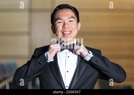 Un giovane che indossa un smoking, regola con cura il suo papillon nero, preparandosi per un evento formale con un sorriso brillante Foto Stock