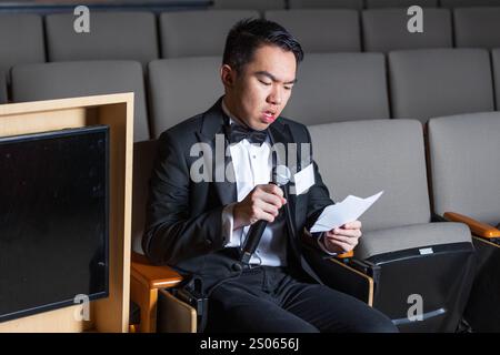 Un giovane studente universitario è a un concorso di lingua pubblica, seduto in una sala da teatro, tenendo le sue schede di scrittura e un microfono, preparando f Foto Stock