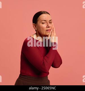 Una donna anziana in rosso preme la guancia con entrambe le mani, mostrando mal di denti o dolore facciale, su un morbido sfondo rosa. Foto Stock
