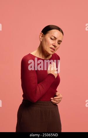Donna anziana, pensionessa si aggrappa al petto in disagio, ritraendo dolore cardiaco o mancanza di respiro sullo sfondo rosa dello studio. Foto Stock
