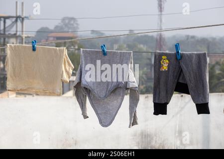 I vestiti bagnati per bambini sono appesi su una linea di abbigliamento con clothespin su un tetto, asciugando al sole e al vento. Foto Stock