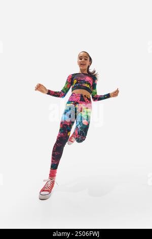 Ragazza vestita con abbigliamento sportivo multicolore astratto, trainer, che salta entusiasticamente in avanti con grande energia su sfondo bianco da studio. Foto Stock