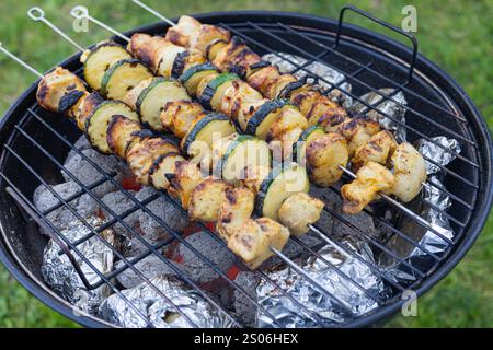 Spiedini di pollo alla griglia e zucchine cucinati su un barbecue a carbone con patate al forno avvolte in un foglio di alluminio. Cucina all'aperto e picnic estivo Foto Stock
