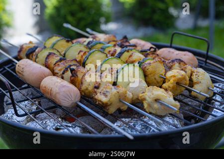 Primo piano di spiedini con pollo alla griglia, zucchine e salsicce su un barbecue a carbone in un giardino lussureggiante. Cucina all'aperto e riunioni estive Foto Stock