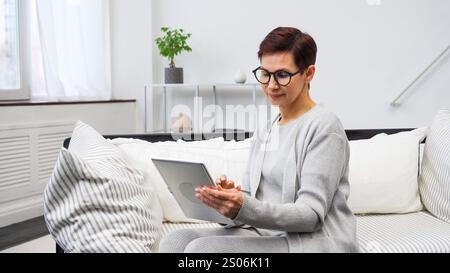 Donna matura con occhiali che reggono un tablet che naviga su app per lo shopping online comodamente seduto sul divano Foto Stock