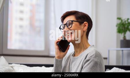 Una donna allegra ride gioiosa mentre parla sul suo smartphone, seduto su un divano in un ambiente rilassato Foto Stock