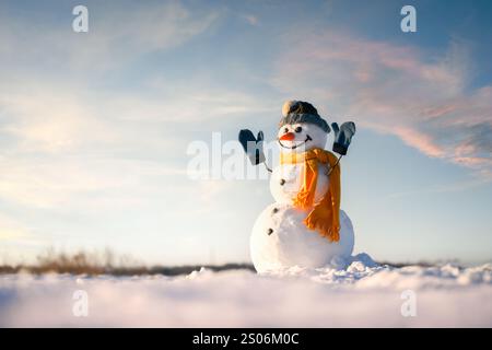 Smiley pupazzo di neve con cappello a maglia e sciarpa gialla con le mani in alto su un campo innevato. Caldo cielo al tramonto con nuvole sullo sfondo Foto Stock