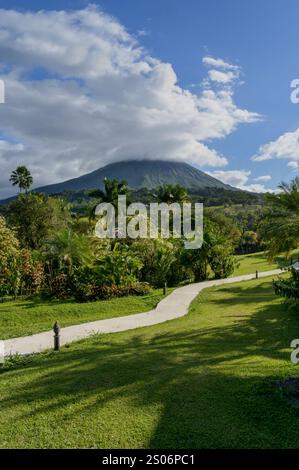 Un percorso circondato da vegetazione tropicale che conduce attraverso l'erba verde con il vulcano Arenal sullo sfondo. Foto Stock