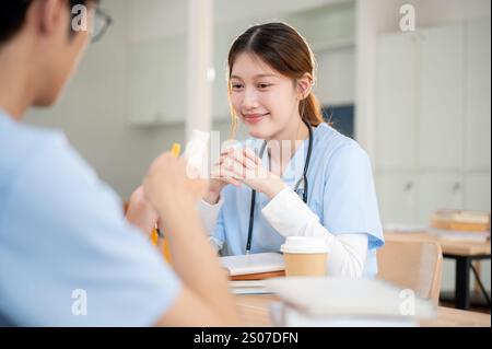 Una positiva, sorridente asiatica studentessa di medicina in scrub sta studiando con il suo compagno di classe maschile in biblioteca, discutendo di ricerca o preparandosi per un Foto Stock