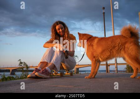 Una giovane donna con i capelli ricci siede su uno skateboard, tiene un guinzaglio e interagisce con il suo cane careliano vicino a un fiume al tramonto, creando una scena tranquilla e avvolgente Foto Stock