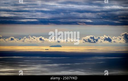 Un tranquillo paesaggio marino caratterizzato da una nave lontana su acque calme sotto un cielo spettacolare pieno di nuvole. La luce del sole si riflette sull'acqua, creando un gol Foto Stock
