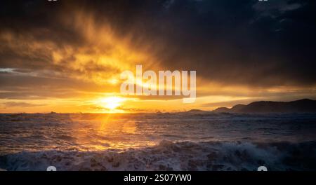 Un tramonto spettacolare sulle turbolente onde oceaniche, con raggi dorati che penetrano attraverso nuvole scure. La scena cattura la bellezza e il potere della natura. Foto Stock