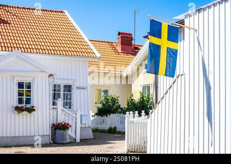 Un vicolo acciottolato si snoda tra le vecchie case in legno di Smögen, Svezia, con una bandiera svedese appesa su un muro di pannelli in legno in una soleggiata giornata estiva. Foto Stock