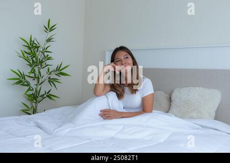 graziosa donna latina su un letto bianco con morbidi cuscini in una camera tranquilla che guarda alla finestra e si gode un momento di relax con un pla verde Foto Stock