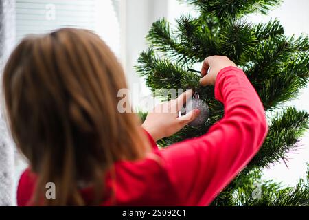 Ragazza che appende con cura l'ornamento della palla glitter argentata sul ramo dell'albero di natale. Foto Stock