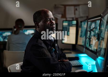 Serio agente di sicurezza afroamericano che si concentra su video CCTV sullo schermo del computer mentre è seduto accanto al posto di lavoro nella sala di osservazione Foto Stock