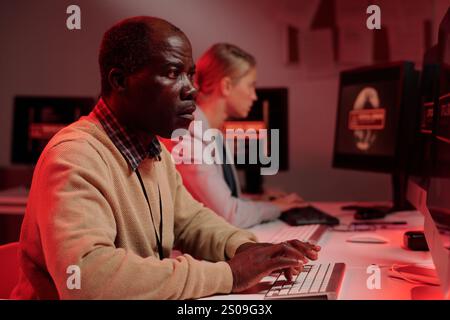 Vista laterale di un serio e maturo agente di sicurezza che cerca di risolvere un problema tecnico mentre si è seduti davanti allo schermo del computer Foto Stock