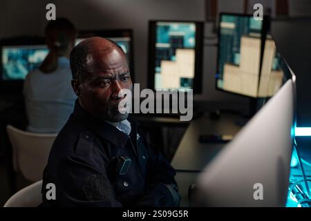 Guardia di sicurezza matura e seria seduta accanto al posto di lavoro nella sala di osservazione e guardando uno degli schermi dei computer desktop Foto Stock