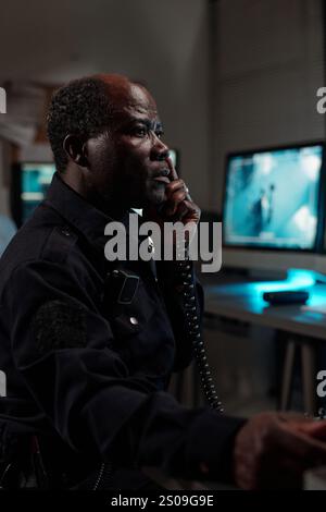 Uomo serio e maturo in uniforme nera della guardia di sicurezza seduto accanto al posto di lavoro nella sala di osservazione e che parla nel ricevitore del telefono Foto Stock