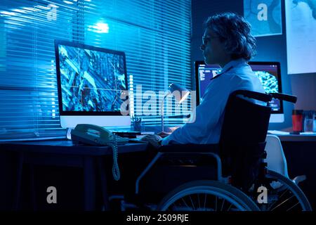 Donna anziana con disabilità seduta in sedia a rotelle di fronte al computer desktop e che guarda i dati grafici Foto Stock